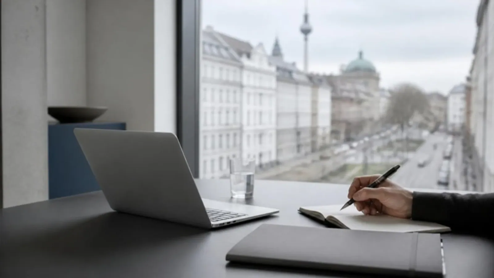 A laptop, notebook and pen on a desk by a window overlooking a Berlin street with the TV tower in the distance