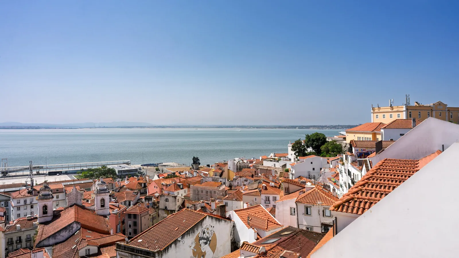 Terracotta rooftops of Lisbon's Alfama district sloping down toward the Tagus River, with church towers on the skyline